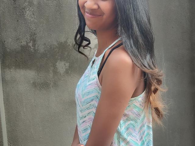 LoderFist has a light brown, smooth skin tone and long, wavy black hair with subtle brown highlights. She's wearing a sleeveless, pastel-colored, chevron-patterned top with thin black straps. She's smiling slightly, showing a small portion of her upper teeth. The top has a soft texture and the chevron pattern includes shades of pink, blue, and green. Her right arm is slightly forward, and the background is a simple, textured, gray wall.