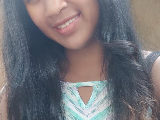 Close-up photo of LoderFist, a woman with long, straight black hair, smiling slightly. She has medium brown skin and is wearing a sleeveless, green and white patterned top with a keyhole cutout. Her lips are pink and she has white teeth. The image is focused on her face and upper torso, with her hair framing her face on both sides. The texture of her hair is smooth and shiny.