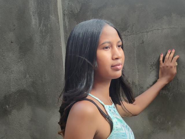 LoderFist is a young woman with medium brown skin and long, straight black hair. She's wearing a light blue patterned tank top. Her right hand, with pink nail polish, is touching a rough, dark gray concrete wall. She's looking to the side with a subtle, confident smile. The light highlights her smooth skin and natural makeup. The composition is simple, focusing on her upper body and the textured wall. The image is sharp with realistic colors.
