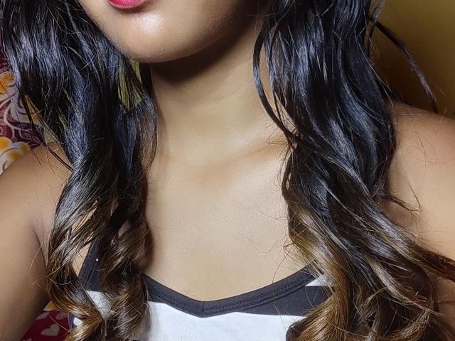 Close-up of LoderFist's face and upper chest. LoderFist has long, wavy black hair with brown highlights, parted in the middle. LoderFist's lips are painted red. She's wearing a white tank top with black trim. The skin tone is medium brown. The hair has a glossy texture. The background is blurred and indistinct.