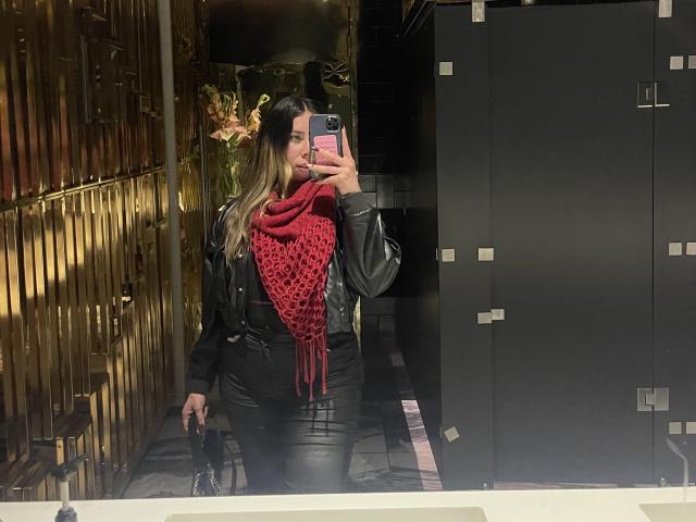 JennyLawry with long brown hair, black leather jacket, red crocheted scarf, black leather pants, holding phone, taking mirror selfie in dimly lit room, holding black purse.