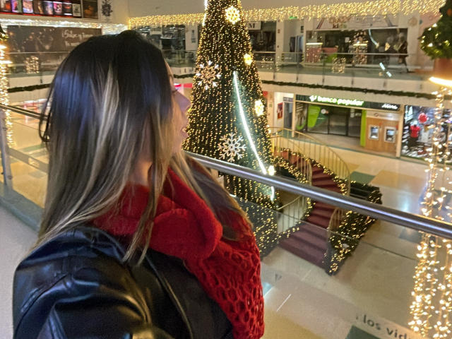JennyLawry with blonde hair, leather jacket, and red knit scarf, looking at a tall, glittery Christmas tree in a mall, surrounded by warm, yellow string lights.
