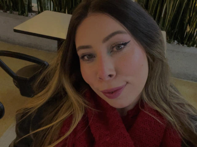 JennyLawry, com cabelos castanhos longos, vestindo um suéter vermelho, tem cílios grossos, batom rosa e um leve sorriso, sentada a uma mesa com uma cadeira e uma luminária..