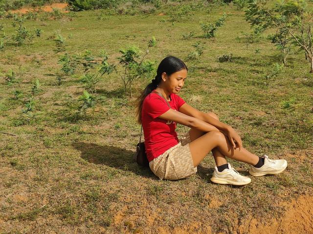 LoderFist sits on a grassy, slightly browned field with small green plants. She has dark, straight hair, wearing a red t-shirt, beige patterned shorts, and white sneakers. Her arms are folded over her knees, and she looks down with a slight smile. Her skin is medium brown. The sunlight casts a shadow to her left. The field has a mix of green and brown, with scattered plants. The overall composition is relaxed and natural.
