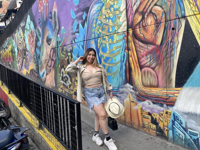 JennyLawry with big ass, tits in a beige crop top, denim shorts, white sneakers, holding a hat, standing in front of colorful graffiti, wearing a cap, smiling.