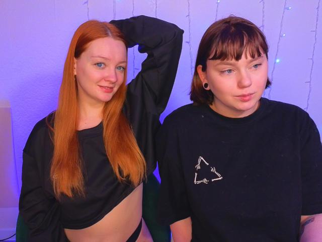 RadiantStwo with red hair, small tits, black shirt, and arm raised, stands next to RadiantStwo with brown hair, black shirt, and star print, both looking at camera.