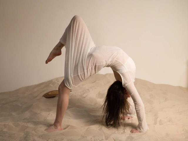 LuluXberry is bending over in a white dress, with her tits pressed against the sand, her ass up, and her head between her legs, in a yoga-like pose.