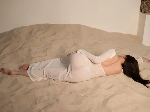 LuluXberry is lying face down on a sand-covered surface, wearing a white, long-sleeve dress that exposes her back and butt. Her arms are stretched out, and her legs are spread.