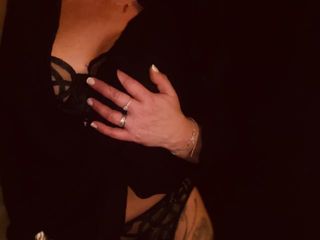 CoquineX69's hand with a silver ring and manicured nails touches her tits, wearing a black lace bra and jacket, under dim lighting.