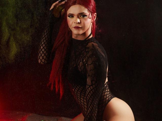Shaina Monroe, con i capelli rossi, indossa un body nero a rete, mostrando il seno e i glutei, in piedi con uno sguardo seducente, sfondo scuro con luci rosse e verdi..