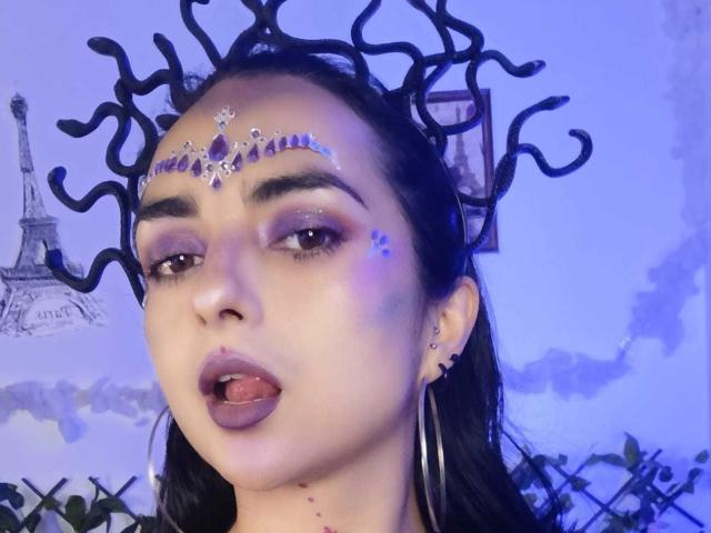 CandyParis with dark hair, wearing a jeweled forehead piece, hoop earrings, and purple eyeshadow, tongue slightly out, looking at camera. Black, curly, abstract hairpiece in background.