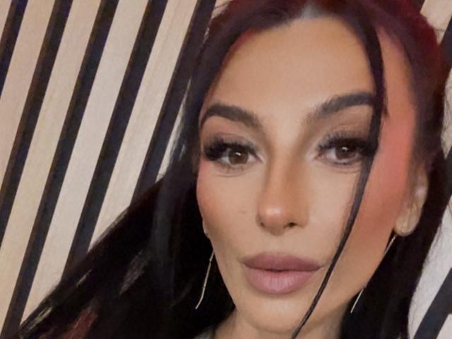 KinkyAmelie has dark, straight hair, brown eyes, and full pink lips. She's wearing subtle makeup and small hoop earrings. Her skin is tan and she looks directly at the camera.
