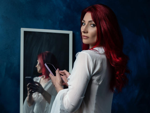 AllyVonD with red hair, white shirt, holding phone, looking at mirror reflection, dark blue background, mirror showing her from side, serious expression, wavy hair, slight smile.