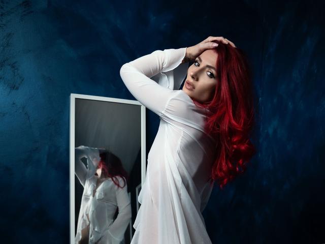 AllyVonD with red hair, white sheer dress, hand in hair, looking back at mirror, reflection shows her ass, dark blue textured wall, soft light, seductive pose.