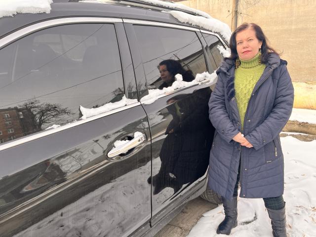 ScarlettWotte, snow-covered black car, green turtleneck, blue puffer coat, black boots, standing, hands together, snow on car roof, reflection in window, winter, snow on ground.