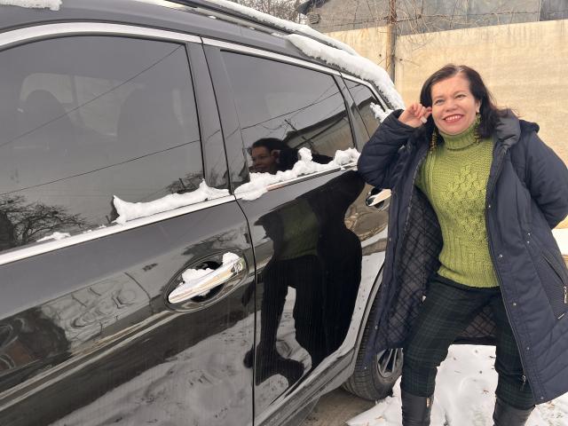 ScarlettWotte stands next to a black car with snow on the roof, wearing a green turtleneck, blue coat, and black plaid pants, smiling with her hand near her face.