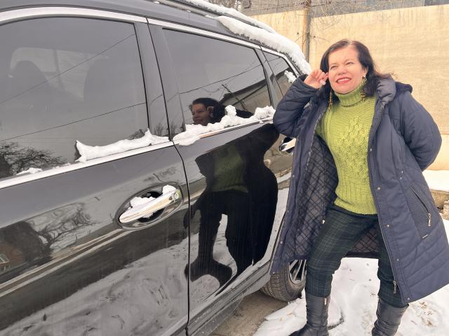 ScarlettWotte with snow on a black car, wearing a green turtleneck, blue quilted coat, and black plaid pants, smiling and touching her face. Snow on roof and ground.