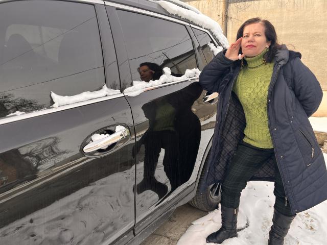 ScarlettWotte with snow on her black car, wearing a green turtleneck, blue quilted coat, and black pants, standing by the door, talking on her phone.