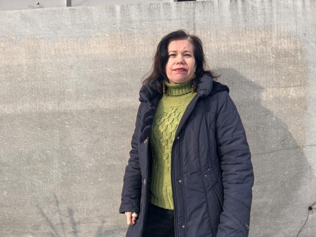 ScarlettWotte, middle-aged woman with green turtleneck sweater, black jacket, and dark hair, standing against a concrete wall, hands in pockets, shadow on her right.