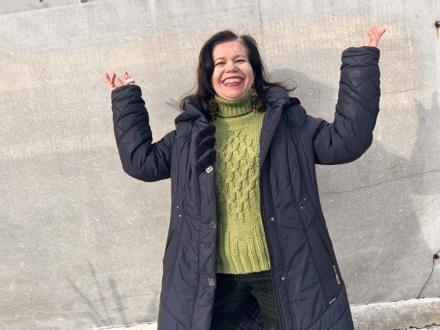ScarlettWotte with arms raised, smiling, wearing a green turtleneck, black puffy coat, and green pants, standing against a concrete wall. Her tits are small, and she's happy.