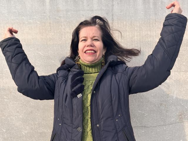 ScarlettWotte with arms raised, smiling, wearing a green turtleneck and black puffy jacket, her brown hair blowing, looking happy and excited, showing her teeth.