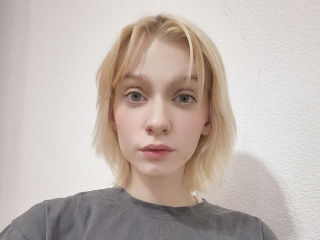 SimonaHeffern has pale skin, blonde messy hair, blue eyes, and pink lips. She's wearing a gray shirt, looking directly at the camera with a neutral expression.