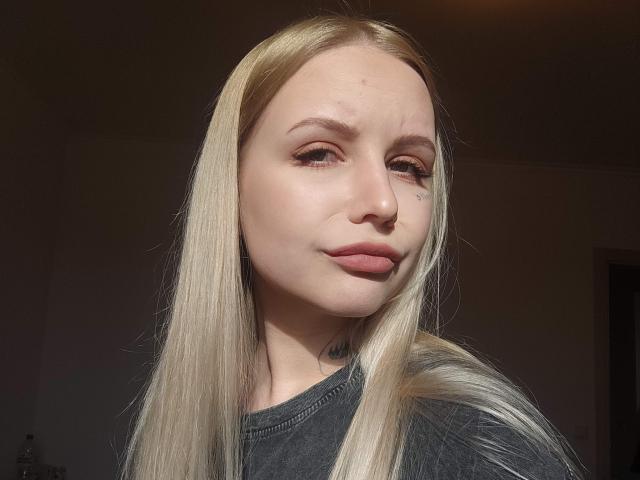 SunshineSweet has long blonde hair, light skin, subtle makeup, pinkish lips, and a small tattoo near her right eye. She's wearing a gray shirt, looking slightly smug.