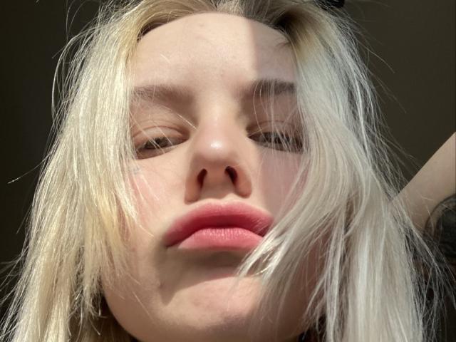 SunshineSweet has blonde, messy hair, pale skin, and pink lips. Her eyes are slightly closed, and she's looking down with a relaxed expression. Her nose is small and upturned.