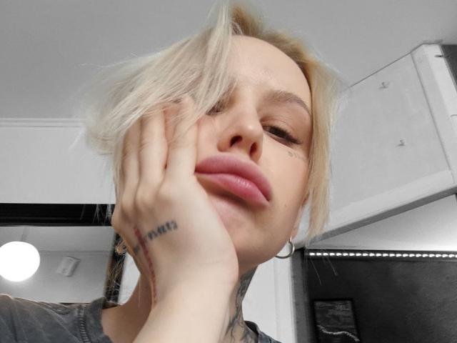 SunshineSweet has blonde hair, pink lips, and a tattoo on her right hand's knuckle. She's wearing hoop earrings, covering her face with her hand, in a selfie.