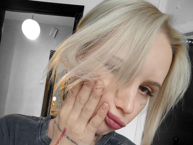 SunshineSweet with blonde hair, partially covering her face with her hand, showing a tattoo on her fingers, wearing a gray shirt, and looking at the camera with a slight smile.