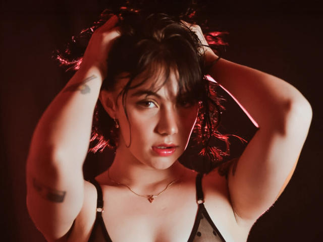 IsabellaArias with hands in messy dark hair, looking down, wearing black sheer top, small tattoos on arms, red lighting, slightly parted lips, small necklace, medium tits, dark background.