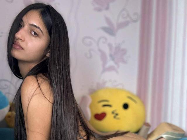 A photo of SugarMaple with long dark hair, looking over her shoulder, showing her ass, in a room with a yellow emoji pillow and floral wallpaper.