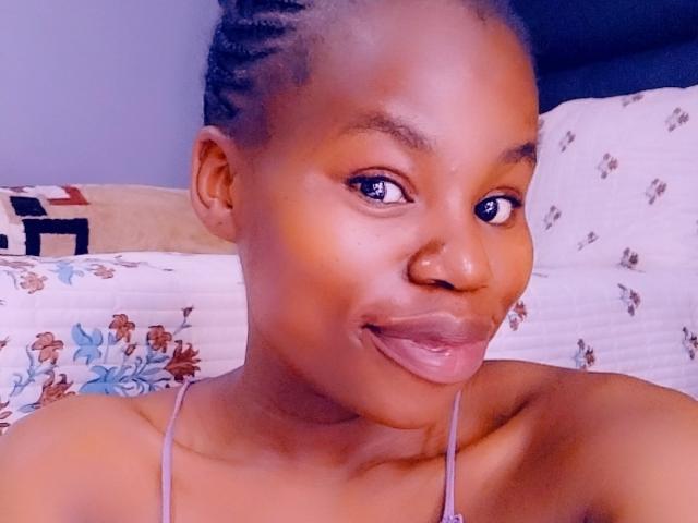 RoundyAss with dark brown skin, braided hair, sticking out tongue, wearing a spaghetti strap top, blue eyes, and a slight smile, in a headshot selfie.
