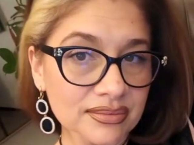 ClassybutNaughty tiene piel clara, ojos marrones y cabello rubio. Lleva gafas negras y aretes redondos con patrones negros y blancos. Sus labios están ligeramente fruncidos..