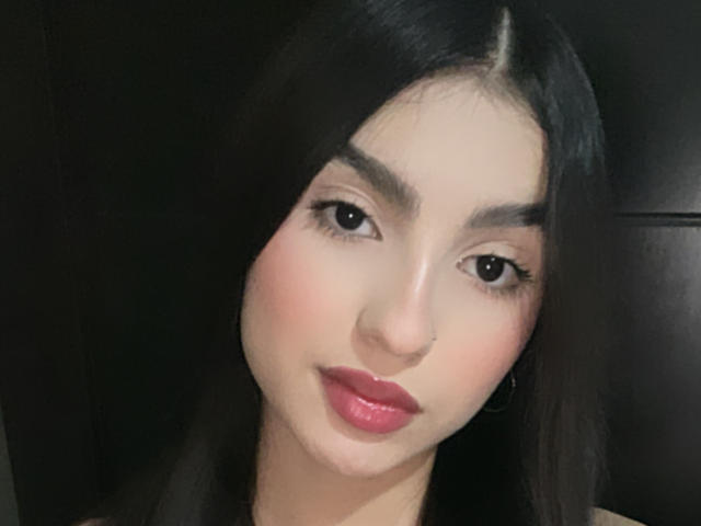MilanMoon has long black hair, brown eyes, and pink lipstick. Her face is slightly tilted with a subtle smile. Her tits are not visible. She's cute.