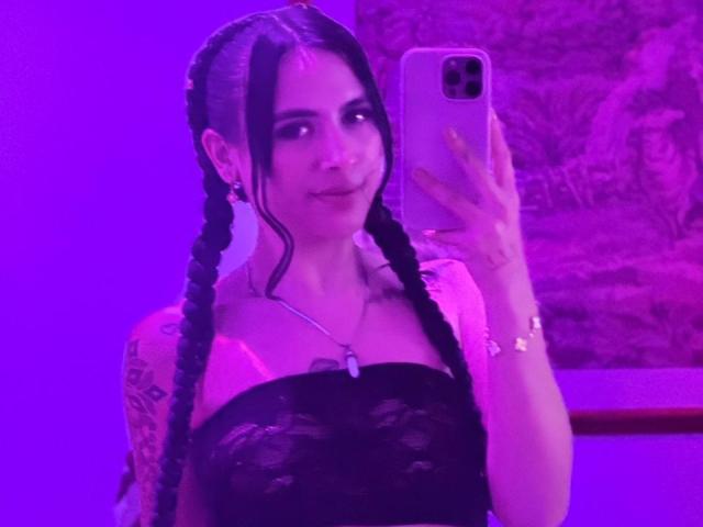 LunaDesire with braided black hair, pink lipstick, and tattoos, taking a selfie in purple lighting, wearing a black lace top, showing her tits, holding a pink phone.