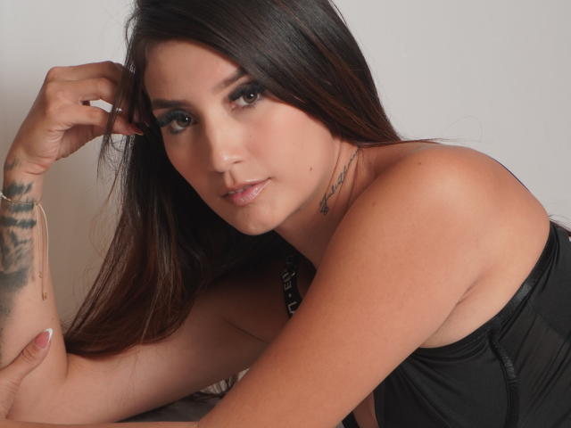 HannahSwan with long brown hair, looking at the camera, has a tattoo on her right forearm, wearing a black strap top, and has glossy pink lips, and brown eyes.