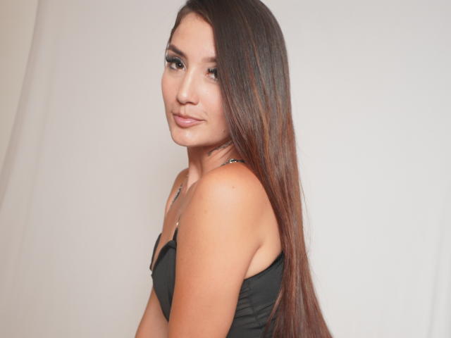 HannahSwan with long brown hair, black tank top, and a cute smile, showing her ass slightly, looking over her shoulder. She has a cute nose and eyeliner.