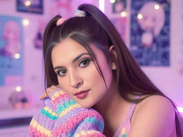 PrinceHermosa with puffy purple lips, wearing a rainbow striped sweater, has long brown hair in pigtails, and is looking at the camera with a cute, sultry expression.