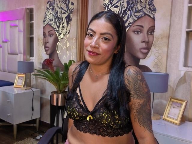 VanessaJohnson with black lace bra, yellow bow, tattooed arm, medium tits, dark hair, red lipstick, standing in room, mirror reflecting her face, potted plant, lamp, framed photo.