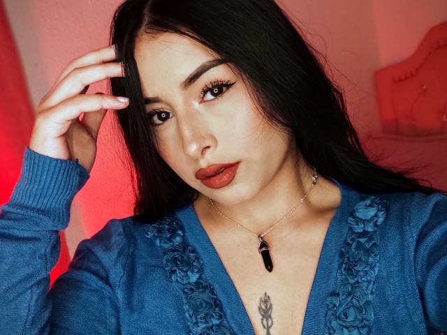 EmilyVv has long black hair, dark red lipstick, blue lace-up sweater, and a black gem necklace. Her hand touches her head, showing a chest tattoo. Red light in background.