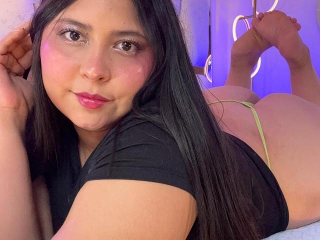 DakotaJhons69, laying on her stomach, black shirt, green thong, butt up, feet in air, black hair, pink blush, pink lipstick, smiling, bare skin, bedroom, purple light.