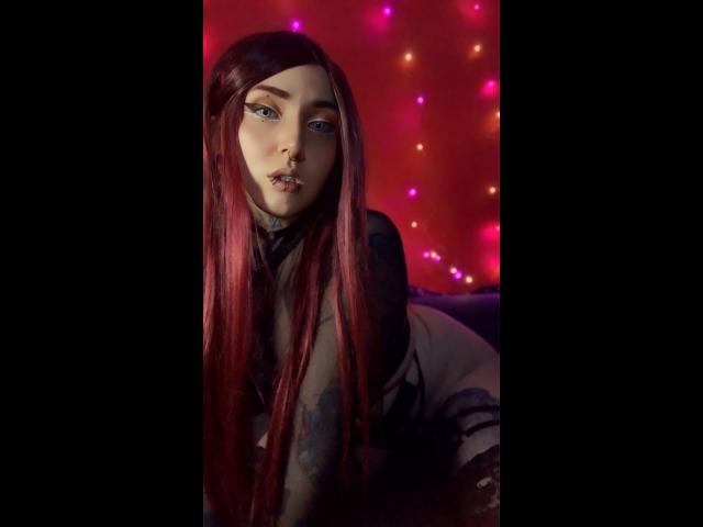 AliceCalloway with long red hair, blue eyes, and tattoos, wearing a black top, looking seductively at the camera, nose ring, and purple lip, in a dark, colorful setting.