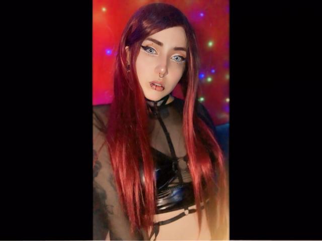 AliceCalloway with long red hair, blue eyes, nose ring, and black leather top, showing tattoos on her arm, and a small nose piercing, looking directly at the camera.