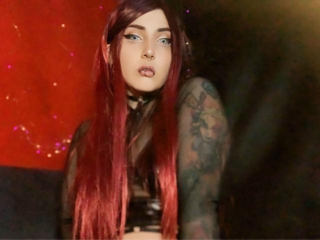 AliceCalloway with long red hair, blue eyes, and a nose ring, tongue out, wearing a black choker and sheer top, tattooed arm, looking seductively at the camera.