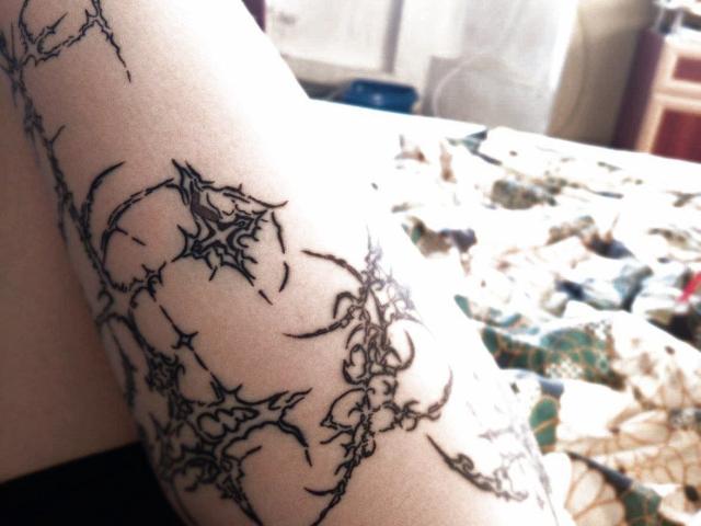 A close-up of NessaV's arm shows intricate black tattoos of twisted, thorny branches. The tattoos are detailed with sharp lines and pointy tips, creating a chaotic yet elegant pattern. The skin is light, and the arm is slightly angled. The background is blurred, with a patterned surface and indistinct objects. The tattoo's dark lines contrast sharply with NessaV's pale skin. The image focuses on the tattoo's complexity and texture.