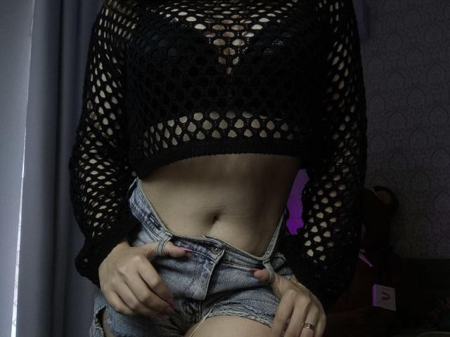 Dreia indossa una maglia nera in rete che lascia intravedere i suoi seni, con l'addome scoperto e le mani che slacciano i suoi pantaloncini di jeans. La sua pelle è chiara e le sue unghie sono rosa..