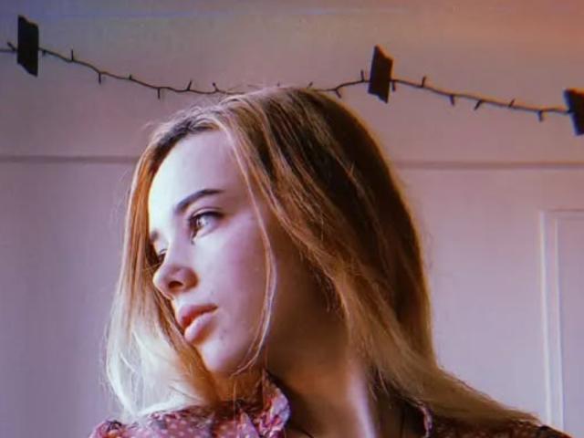 EvieEve with blonde hair, pouty lips, and a side profile looking to the left, wearing a floral shirt, string lights in the background.