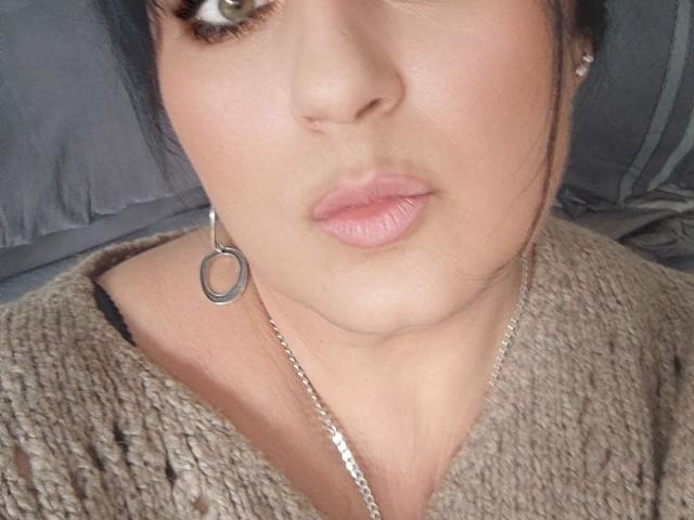BrigitteXLove's face with pouty pink lips, brown eyes, and a silver hoop earring, wearing a brown knit top, showing a bit of her black strap top.