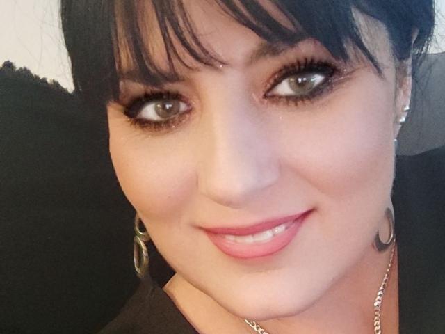 BrigitteXLove has dark brown eyes, black eyeliner, pink lipstick, and a small tattoo on her right cheek. She's smiling with her chin slightly tilted. She's wearing a black top.